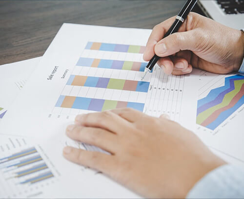 An individual’s hand holds a pen, pointing to a colorful bar graph on a printed paper. Additional graphs and data analysis charts are visible on the desk, conveying a professional setting where actuarial services involving data interpretation and financial analysis are being conducted.