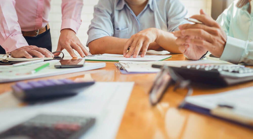 A group of professionals is engaged in a financial analysis, working with documents and a calculator on a table. The focus is on their hands and the papers, indicating a collaborative effort in reviewing detailed financial data.