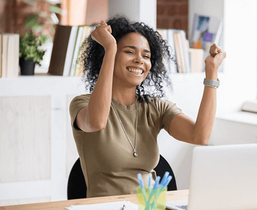 An individual with their arms raised in a gesture of success or celebration sits at a desk in front of a laptop, possibly indicating satisfaction with work, such as the successful management of employee benefits accounting as offered by Prima Consulting. The individual’s face is obscured for privacy.