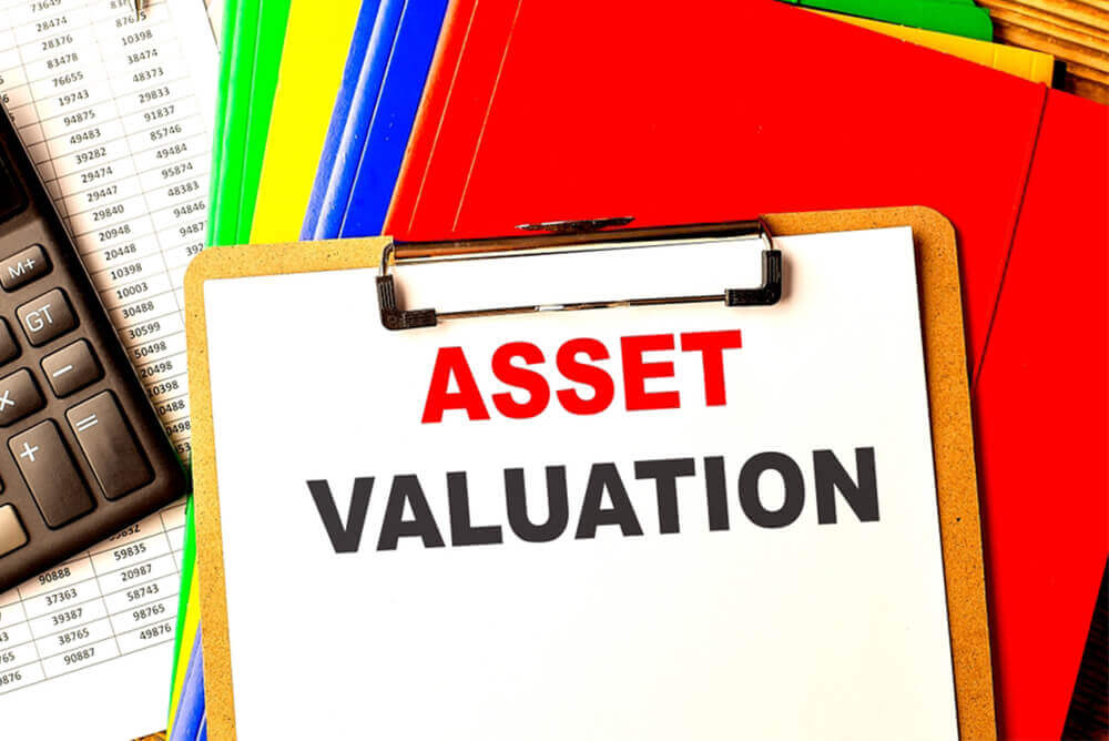 A clipboard with the words “ASSET VALUATION” printed in red on a white paper is prominently displayed in the foreground. Behind it, an array of colorful folders, a calculator, and printed financial documents with numerical data are visible, suggesting a business or accounting setting focused on financial management and asset evaluation.