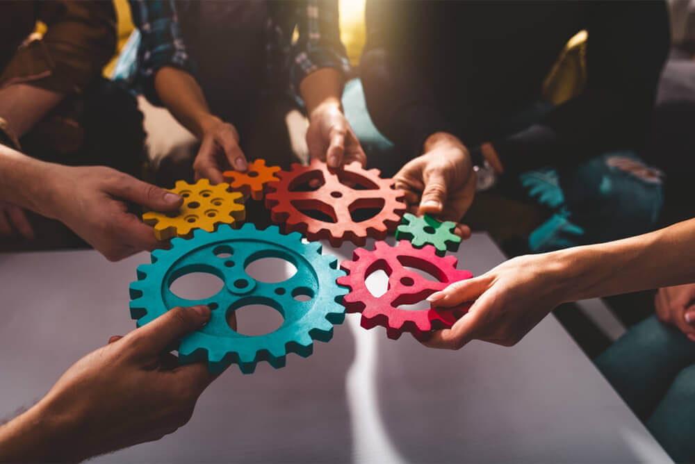 A group of hands coming together, each holding a colorful gear, symbolizing teamwork and integration in a business setting. The gears are interlocking, suggesting the seamless merging of companies or systems as referenced by IFRS 3 Business Combination.