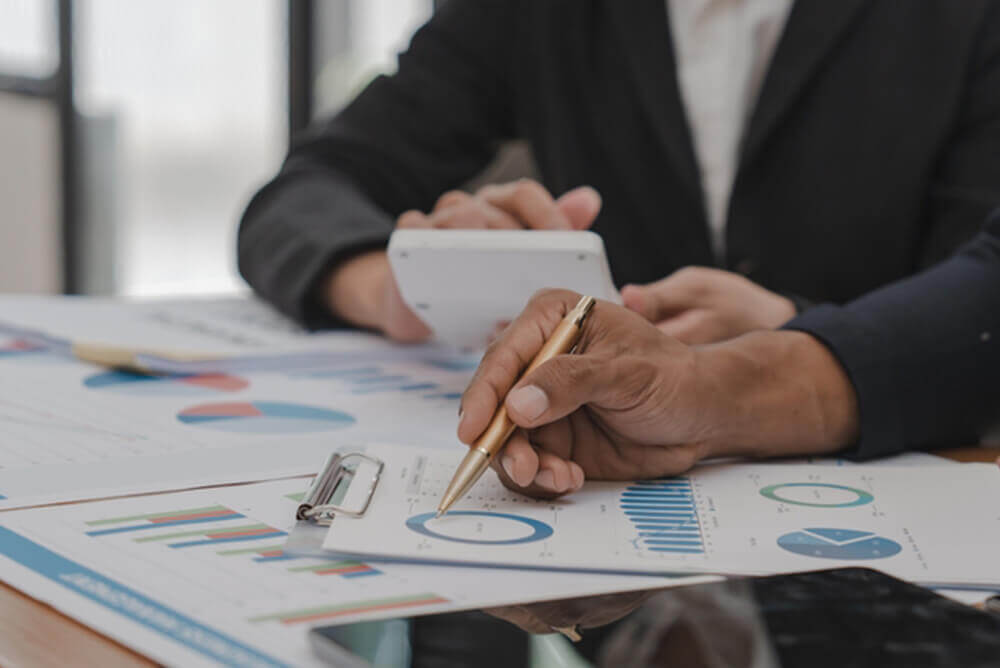 A professional setting featuring a person’s hands, one holding a pen and the other a smartphone, reviewing and working on financial documents with colorful charts and graphs. The focus on the hands and documents highlights the analytical process involved in financial reporting.