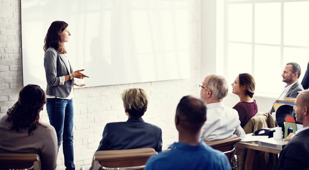 A professional training session in a bright room with large windows. An instructor, facing away from the camera, is gesturing towards a blank projection screen while addressing attendees seated in rows. The attendees are focused on the instructor, suggesting an engaging educational environment.