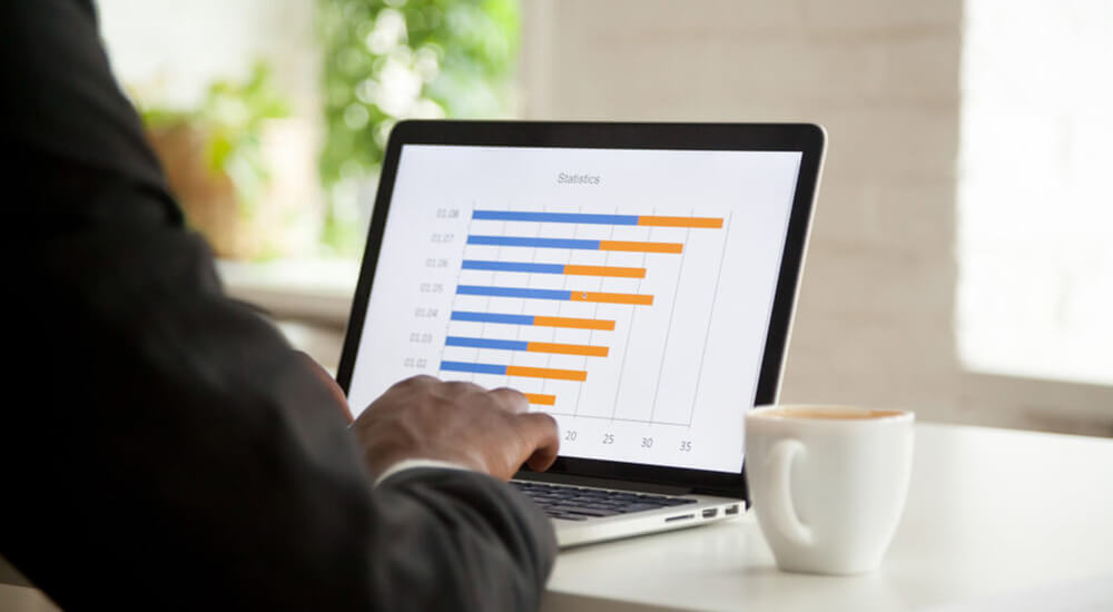 A person is working on a laptop displaying a colorful bar graph with statistical data, likely related to actuarial valuation. The graph shows various bars in shades of blue and orange, indicating different values against a numerical scale. The laptop is on a desk with a blurred background, suggesting an office setting. A cup of coffee sits next to the laptop, indicating a work environment