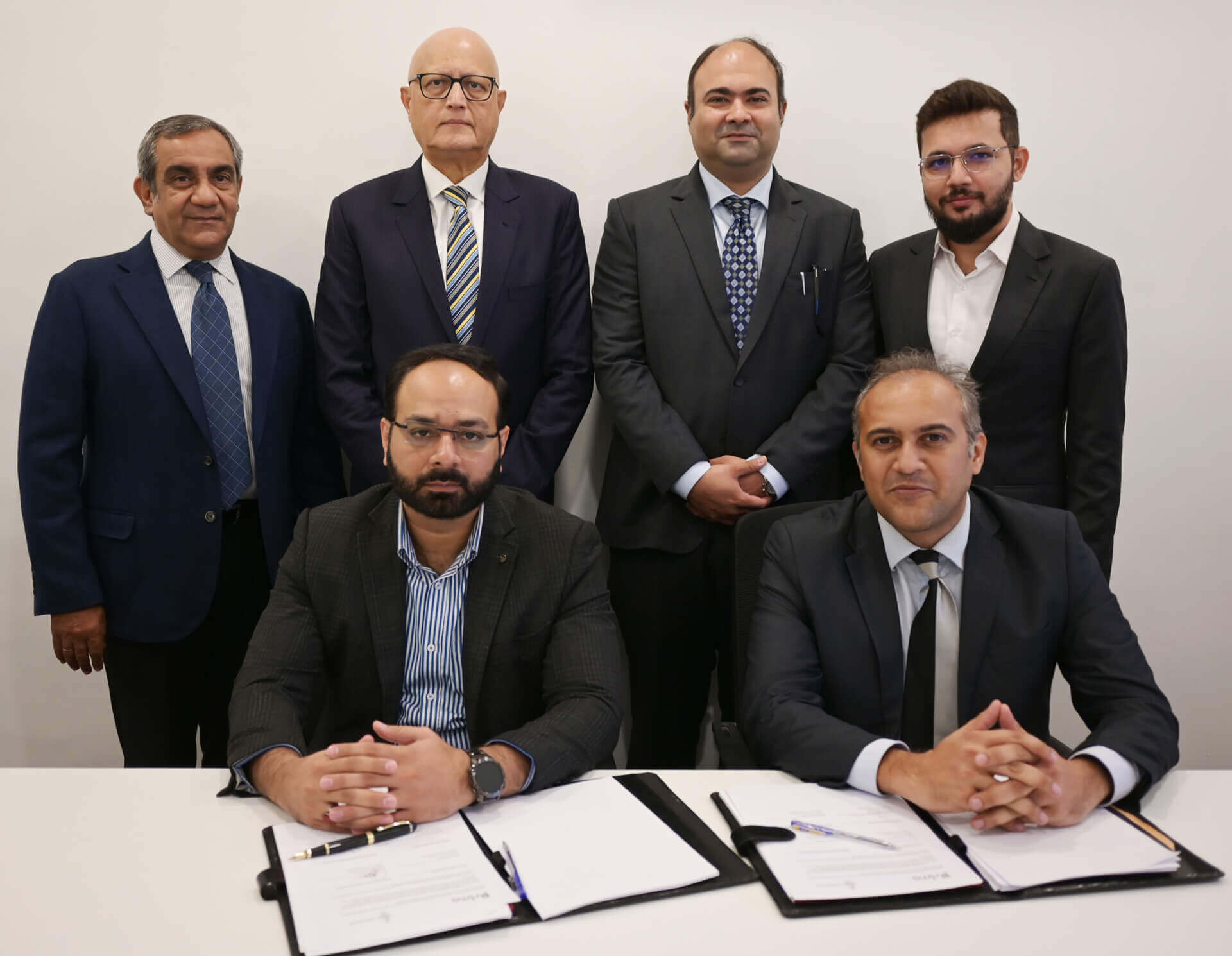 A signing ceremony with two seated individuals at the forefront signing documents and four standing individuals behind them in a professional office setting.