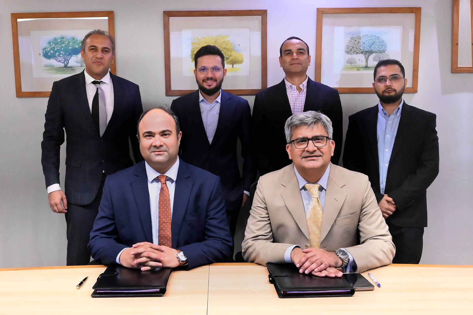 The image captures a formal business setting where two individuals, identified as Faisal Khan (CEO of IGI Insurance) and Ibrahim Zahidie (Director Advisory & Transformation at Prima Consulting), are seated at the forefront with documents on the table, symbolizing a partnership agreement. Behind them stand four professionals: Shabih Ahmed Arif (Director Actuarial Services at Prima Consulting), Sumar Sayani (Director AI & Technology at Prima Consulting), Syed Awais Amjad (CFO of IGI Insurance), and Murtaza Feroz Jandga (Finance Controller at IGI Insurance). All individuals are professionally dressed, and the setting suggests a significant corporate event.