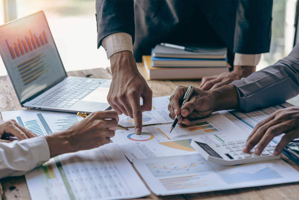 A professional setting with multiple individuals engaged in business planning and analysis. The image shows hands pointing at charts and graphs on paper, indicating a collaborative effort in operational advisory. A laptop displaying additional data charts is also visible, suggesting a data-driven approach to business consulting.