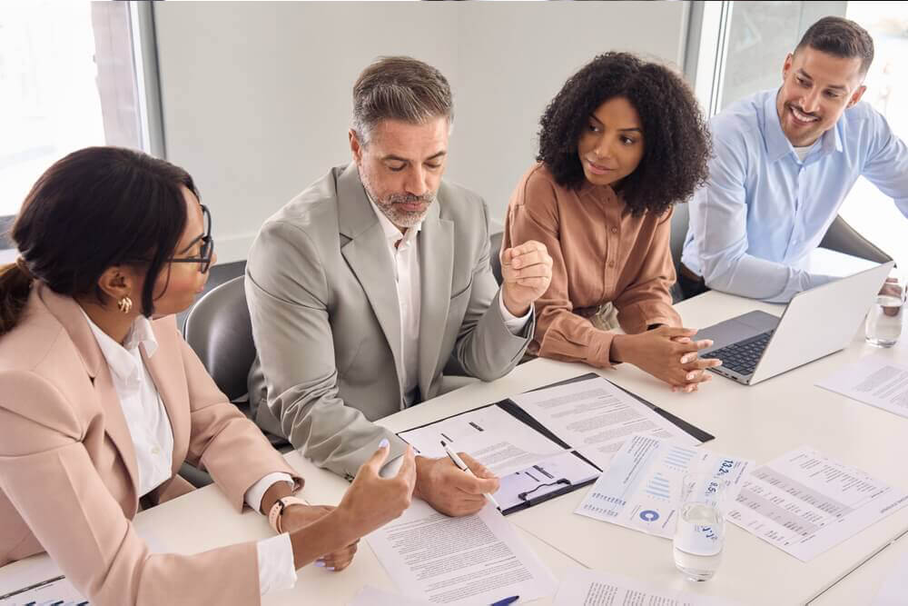 A professional meeting in progress featuring four individuals around a table, with their faces obscured for privacy. One person is taking notes, another is gesturing towards a document, and the others are attentively engaged in the discussion. Visible on the table are papers with graphs and charts, a laptop, and a bottle of water, indicating an analytical or strategic business environment. This image likely represents the collaborative and expert nature of Prima Consulting’s Financial & Risk Advisory Services