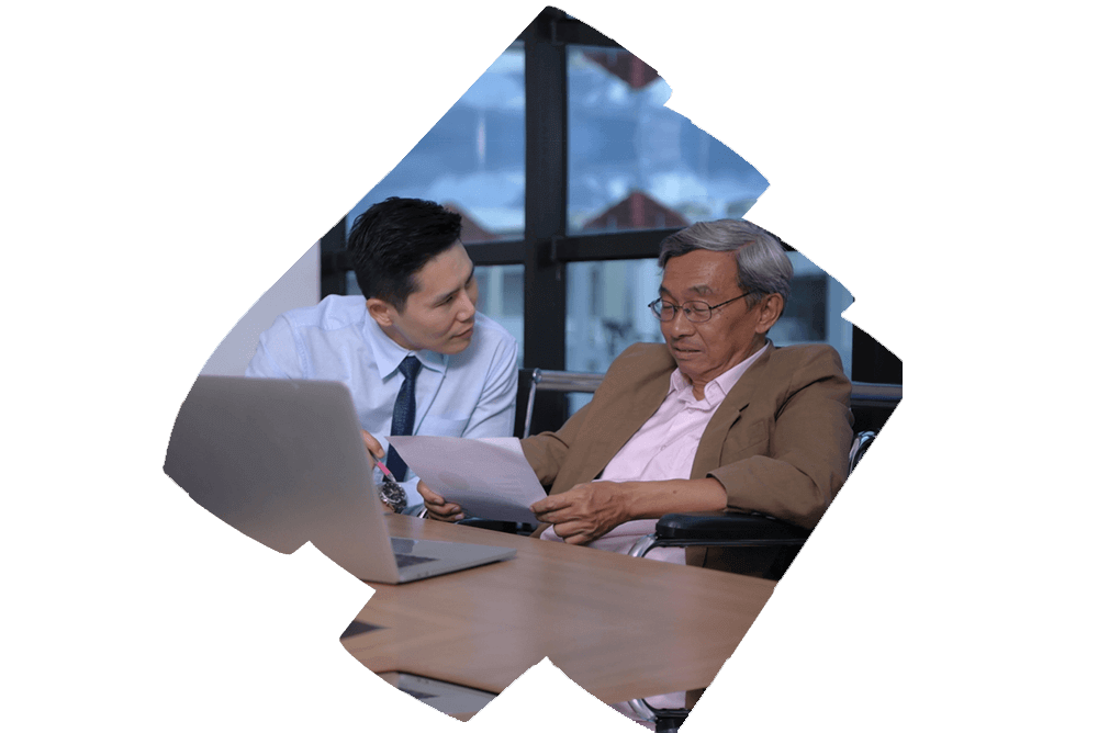 two individuals in a professional setting, one seated in a wheelchair and the other crouched beside them, engaging in what appears to be a serious conversation. Both individuals’ faces are obscured for privacy. The setting suggests a focus on inclusivity within the workplace, aligning with the concept of Benefit Harmonization which aims to standardize employee benefits across an organization.