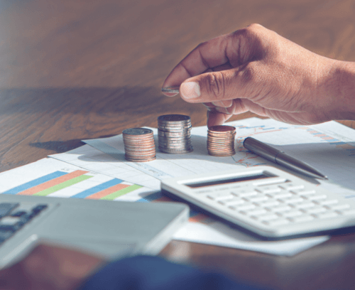 A close-up image of a hand stacking coins in ascending order on a desk with financial charts, a calculator, and additional coins laid out. The scene suggests financial planning or analysis.