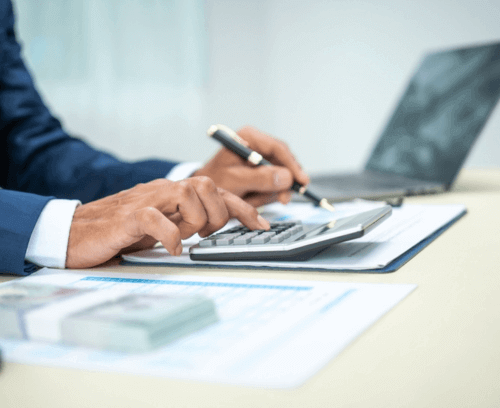 A close-up image of a person’s hands using a calculator and holding a pen, with documents and a laptop in the background, suggesting financial work or analysis.