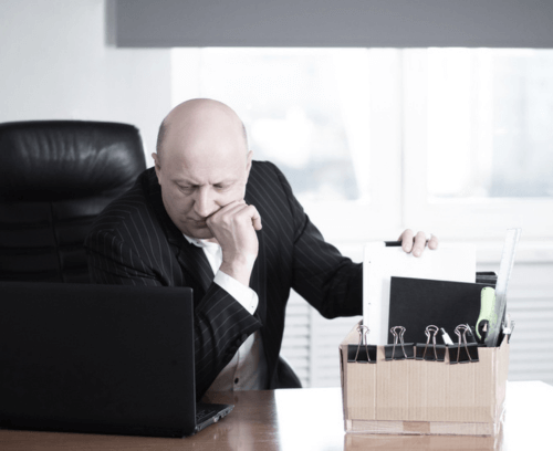 An individual in a business suit is seated at a desk, facing away from the camera, with their face obscured for privacy. The person appears to be in an office setting, holding a document or folder while gesturing with the other hand as if in discussion or explanation. A briefcase and what seems to be a laptop are also visible on the desk, suggesting a professional environment.