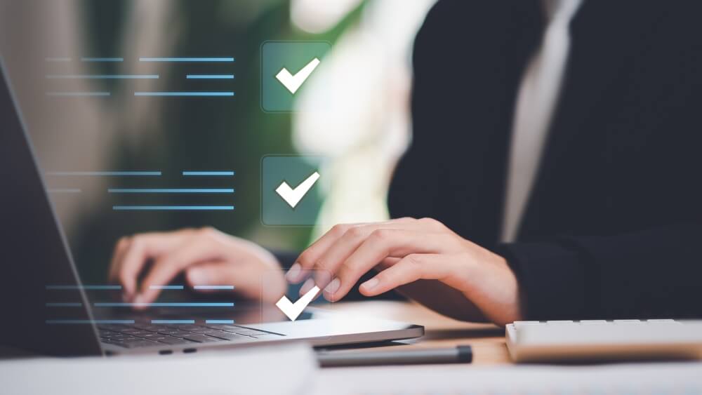 A person is typing on a laptop with a keyboard visible in the foreground. The image features three checkmarks in a vertical line, symbolizing completed tasks or goals. The person is dressed in business attire, suggesting a professional setting.