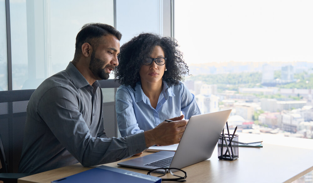 Two individuals are seated at a desk in a modern office with large windows overlooking a cityscape. They are engaged in a discussion while looking at a laptop screen. The desk has office supplies, including a pen holder and a folder.