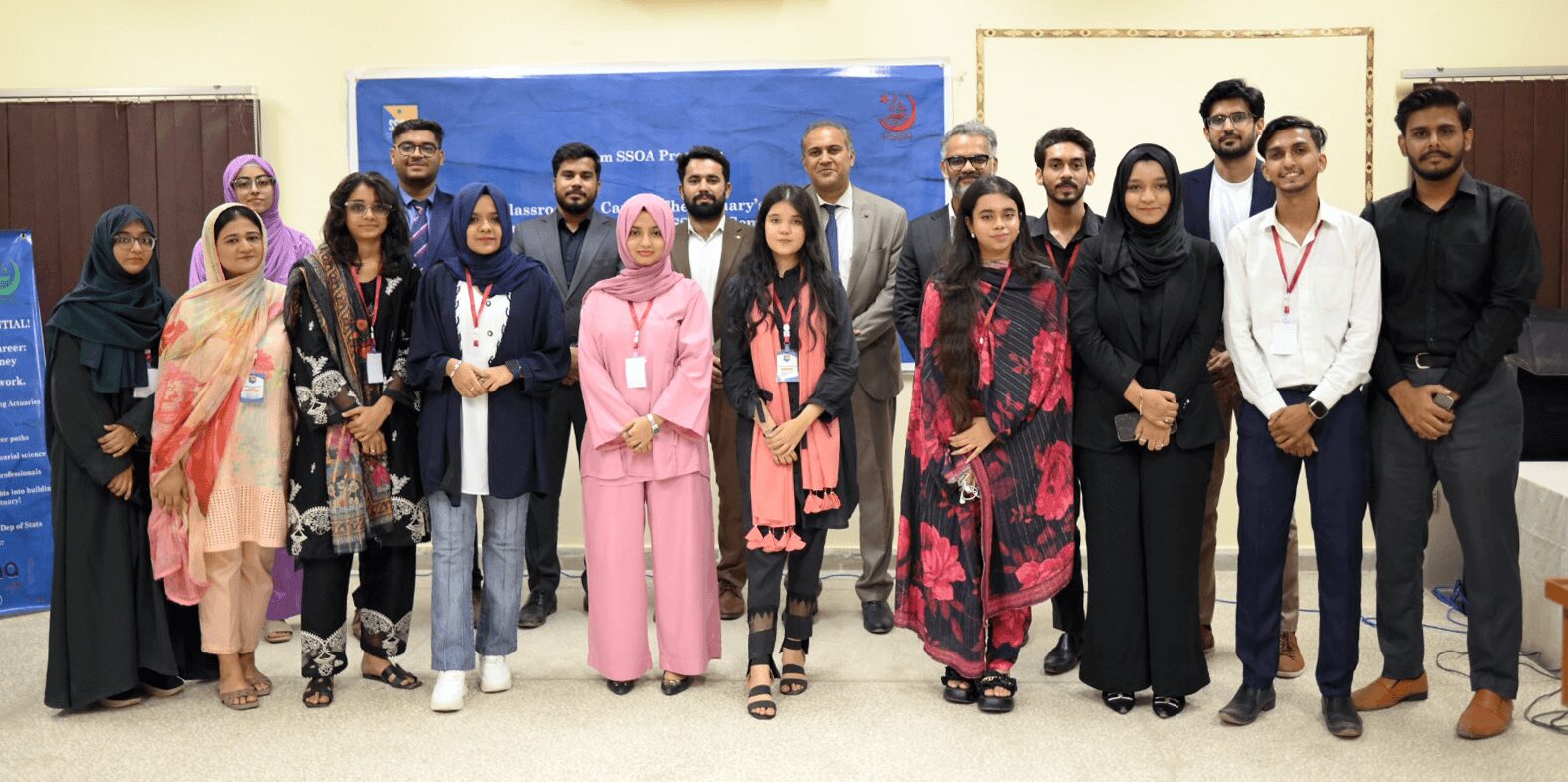 A group of individuals stands together in a room, posing for a group photo. They are dressed in a variety of professional and semi-formal attire, with some wearing traditional clothing. The background features a blue banner and informational posters, indicating an event or gathering.