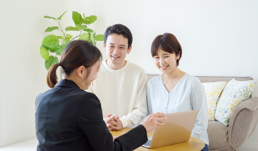 A financial advisor is sitting across from a couple, discussing financial planning and analysis (FP&A) with a laptop on the table. The advisor is pointing at the laptop screen, likely explaining financial strategies and tools to help the couple achieve their financial goals. The setting is a bright, modern living room with a plant and a couch in the background, emphasizing a comfortable and professional environment.