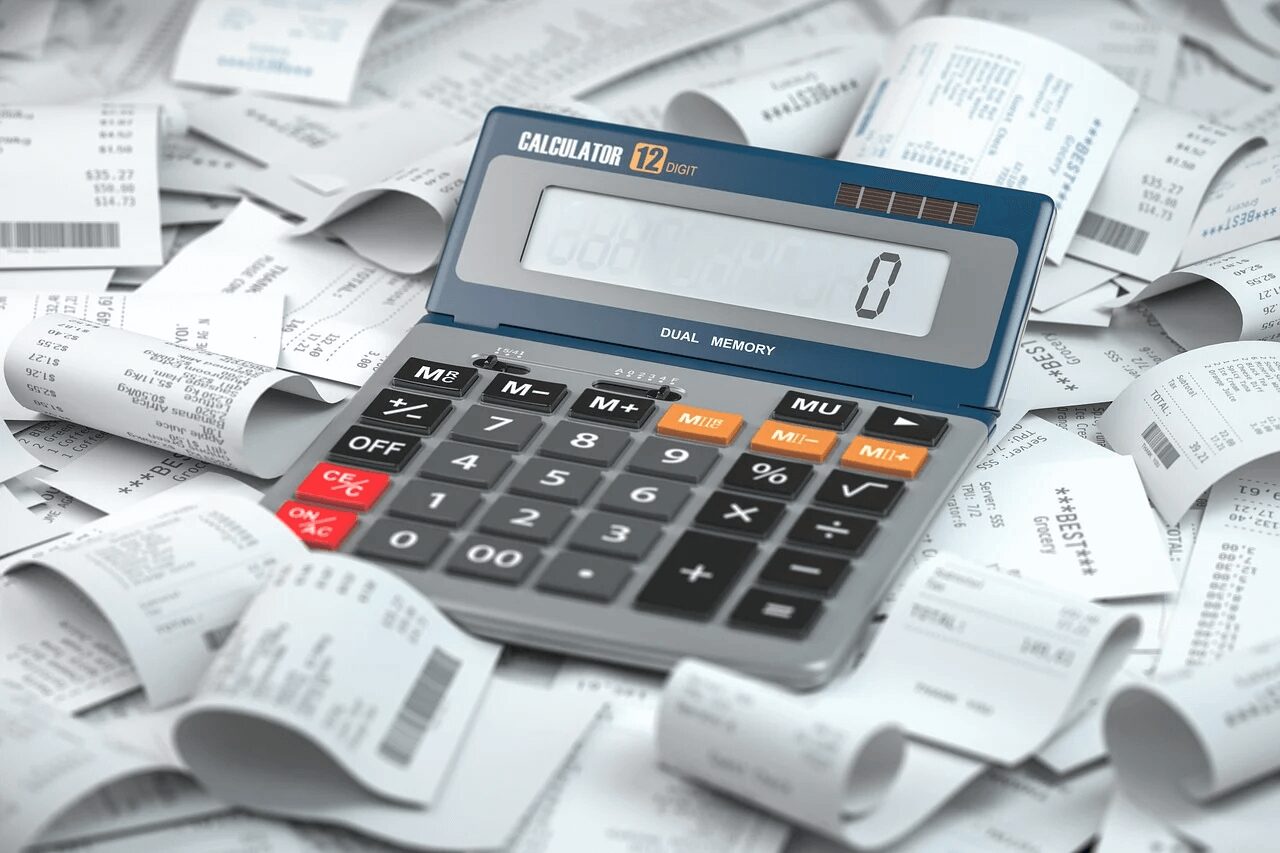 A 12-digit calculator with a "Dual Memory" feature is placed on a pile of various receipts. The calculator display shows the number zero, and the receipts around it contain printed text and numbers, indicating financial transactions.