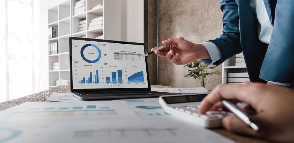 Audit Support: A business meeting scene with two professionals analyzing financial data on a laptop screen. The screen displays various charts and graphs, including a pie chart, bar graphs, and a line graph, indicating financial performance metrics. One person is pointing at the screen with a pen, while the other is using a calculator, emphasizing the collaborative effort in audit support to ensure accurate financial reporting and compliance