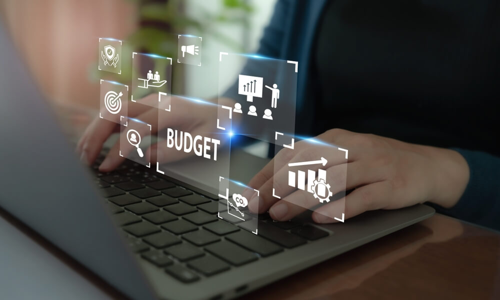 A person types on a laptop with various financial and business-related icons floating above the keyboard, including a target, a graph, and a gear, symbolizing the integration of advanced forecasting tools and predictive analytics for effective budgeting and forecasting.