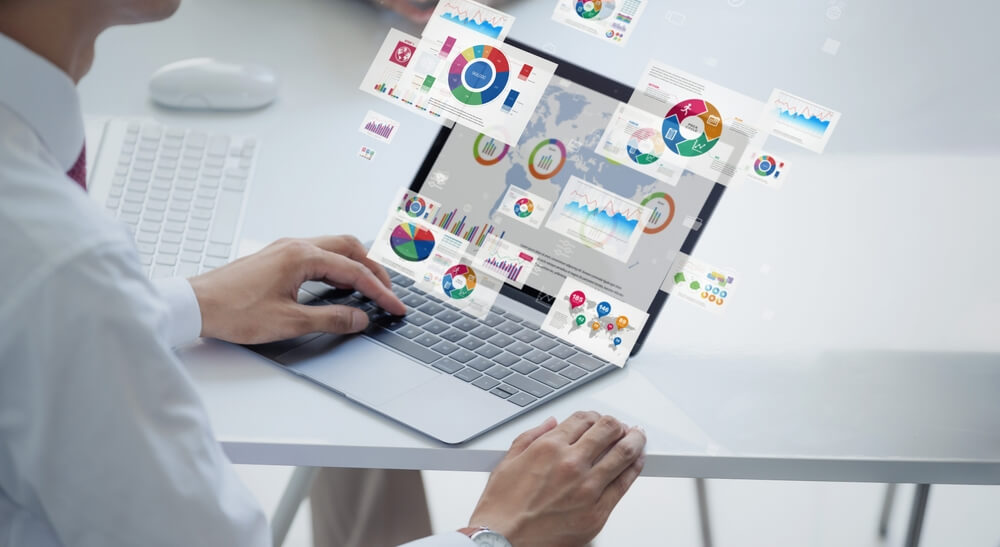 A person is working on a laptop at a desk, with various colorful charts and graphs floating above the laptop screen. These charts include pie charts, bar graphs, and line graphs, indicating data analysis and visualization. The scene suggests the use of advanced software tools for data management and financial analysis and cloud bookkeeping