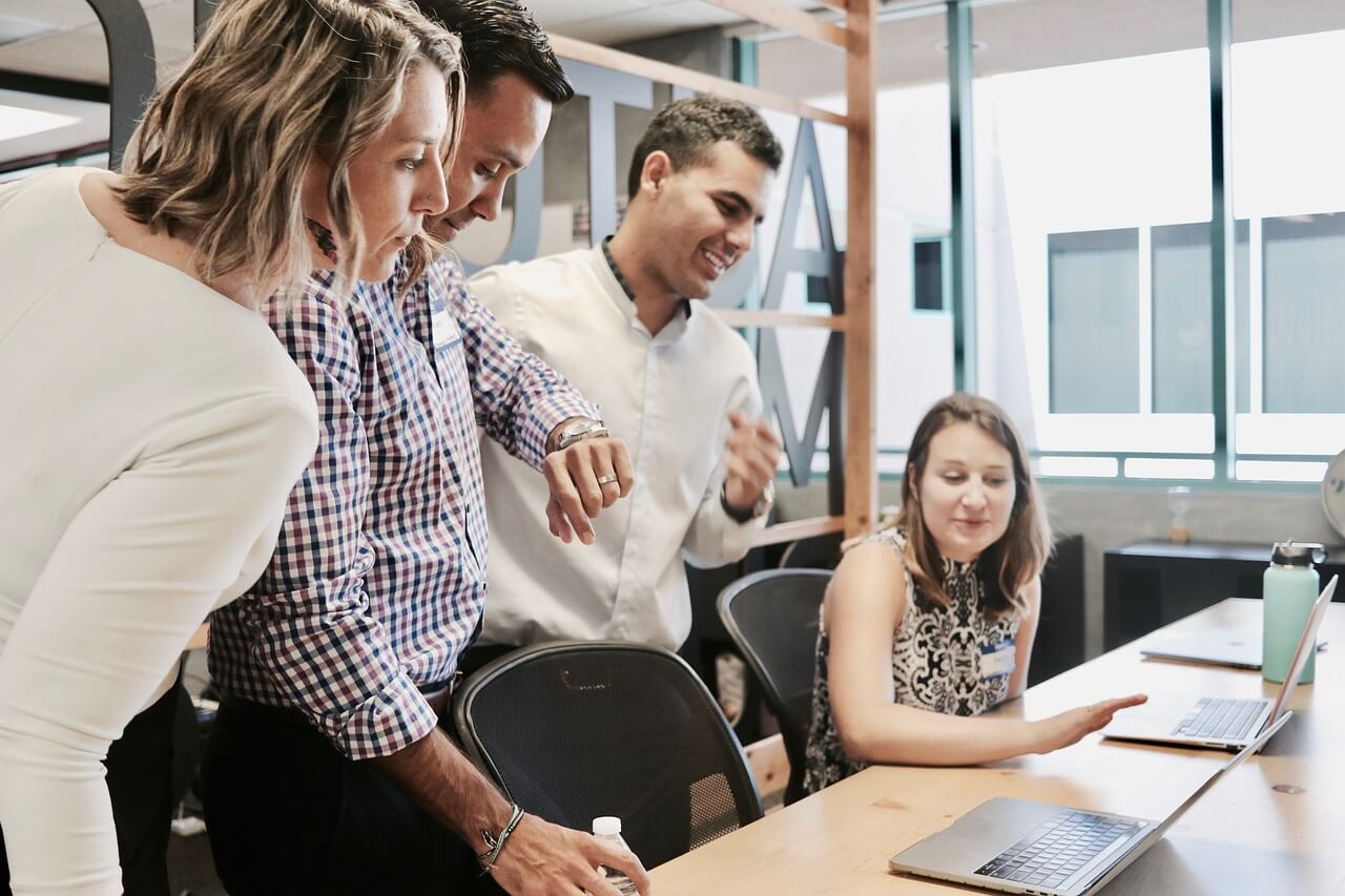 A group of four people are gathered around a desk in an office setting. Three of them are standing and looking at a laptop screen, while one person is seated and pointing at the screen. The office has large windows and modern furniture, creating a collaborative work environment.