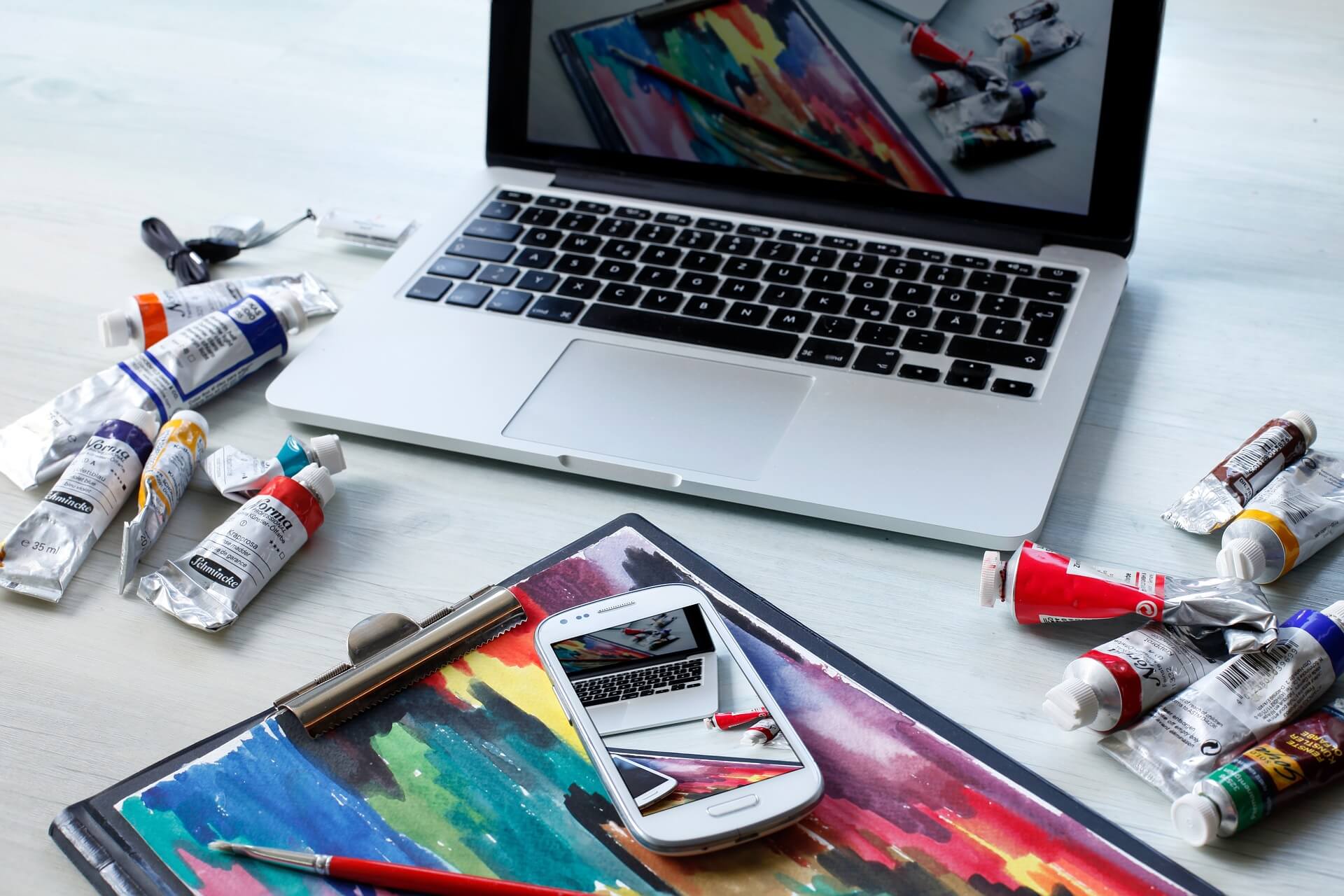 a workspace with a laptop, a smartphone, and various tubes of paint scattered around. The laptop screen displays an image of a colorful painting, and the smartphone, placed on top of a sketchbook with a similar colorful painting, reflects the same image. Paintbrushes and other art supplies are also visible, indicating a creative environment.