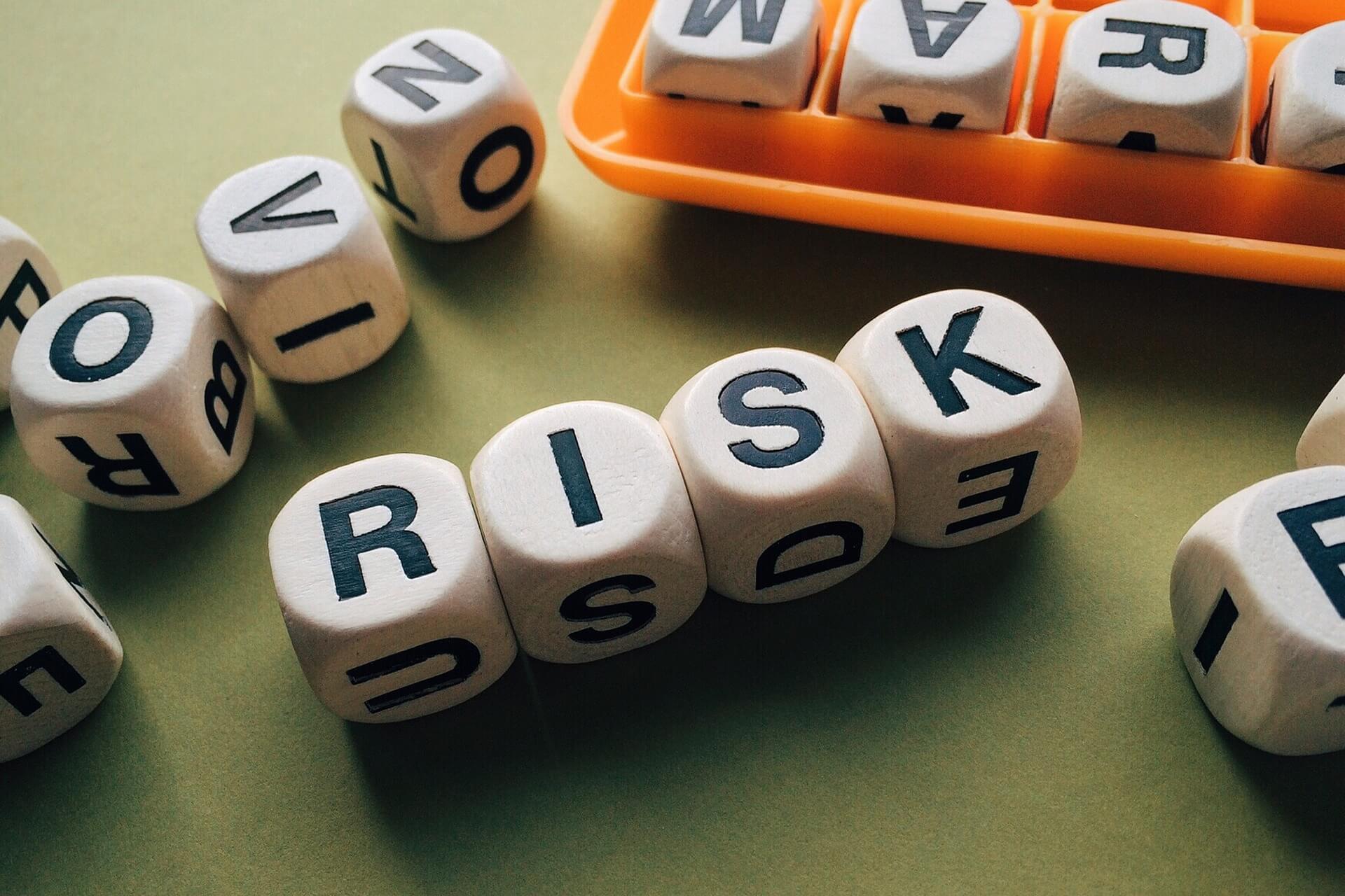 several wooden cubes with black letters on them, arranged to spell the word "RISK." Some cubes are scattered around, and others are in an orange tray. This image is relevant as it visually represents the concept of risk, which ties into Prima Consulting's services focused on risk management and controls.