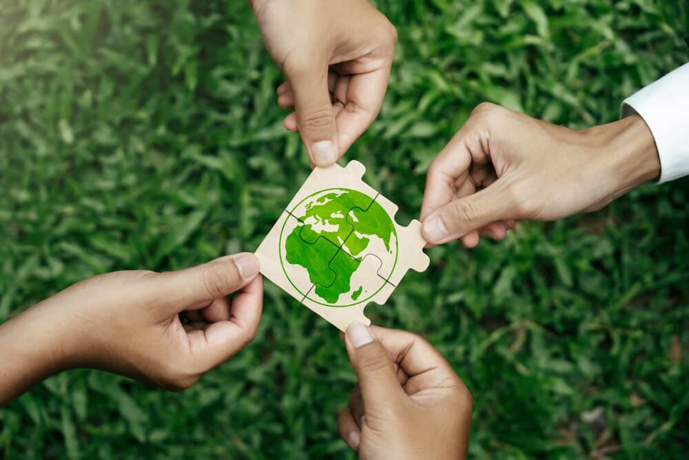 Three hands holding pieces of a puzzle with a green Earth illustration, symbolizing collaboration and sustainability. ESG Consulting Services in KSA, UAE & Pakistan.