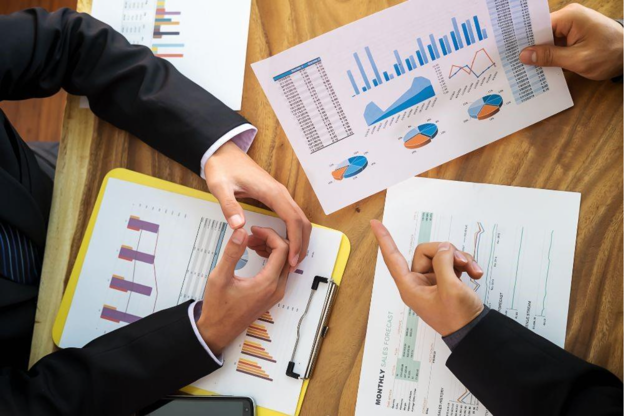 Overhead view of a business meeting with hands pointing at documents and charts on a wooden table, focusing on IFRS 2 compliance analysis. 