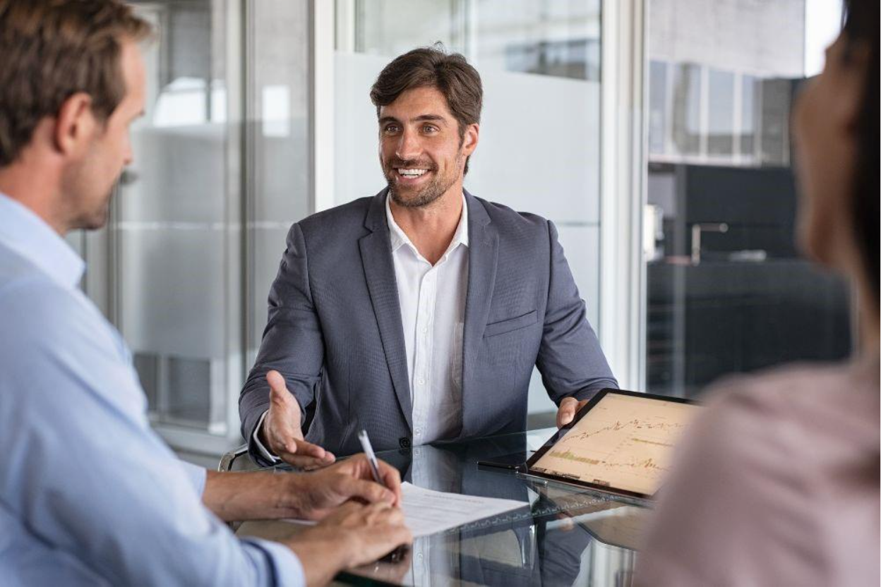 Business meeting discussing IFRS 2 compliance with a professional in a suit gesturing, holding a tablet displaying financial charts, and two colleagues reviewing documents at a glass table. 