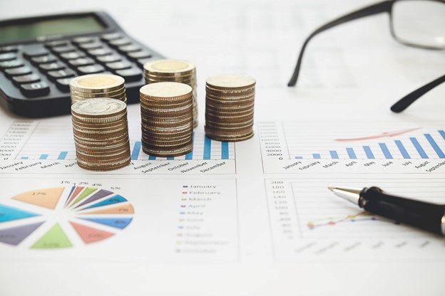 A table with stacks of coins, a calculator, and financial documents displaying actuarial valuation charts, including bar and pie graphs, alongside a pen and glasses.