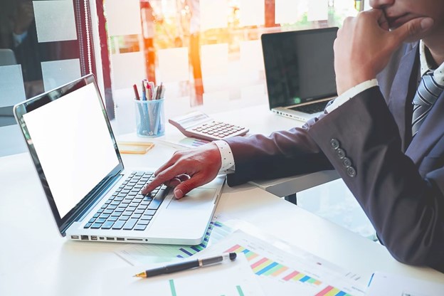 A professional using a laptop with actuarial valuation services charts and documents on the desk in a bright office with large windows.