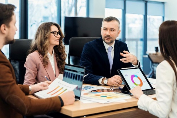 A business meeting with professionals discussing actuarial valuation, surrounded by laptops and tablets showing pie charts and financial documents on the table.