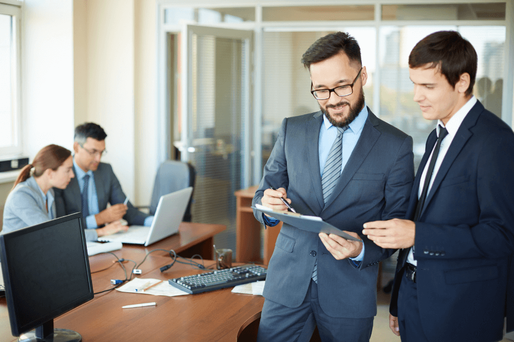 Zwei Männer im Anzug besprechen Dokumente in einem modernen Büro, während im Hintergrund ein weiteres Team arbeitet – professionelle Zusammenarbeit im Bereich Finanzdienstleistungen.
